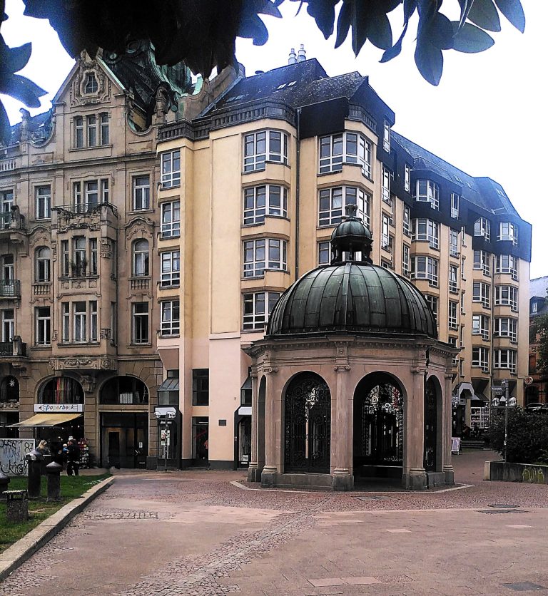 Digitale Zeitreise Wiesbaden- Kranzplatz und Kochbrunnentempel Historisches Gebäude mit einem Rundbau und Kuppel, umgeben von weiteren Architektur.