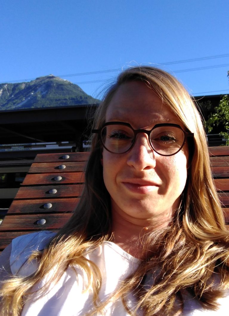 Erkundung und Abenteuerlust Junge Frau mit Brille sitzt auf einer Holzbank vor einem Berg und klarem Himmel.