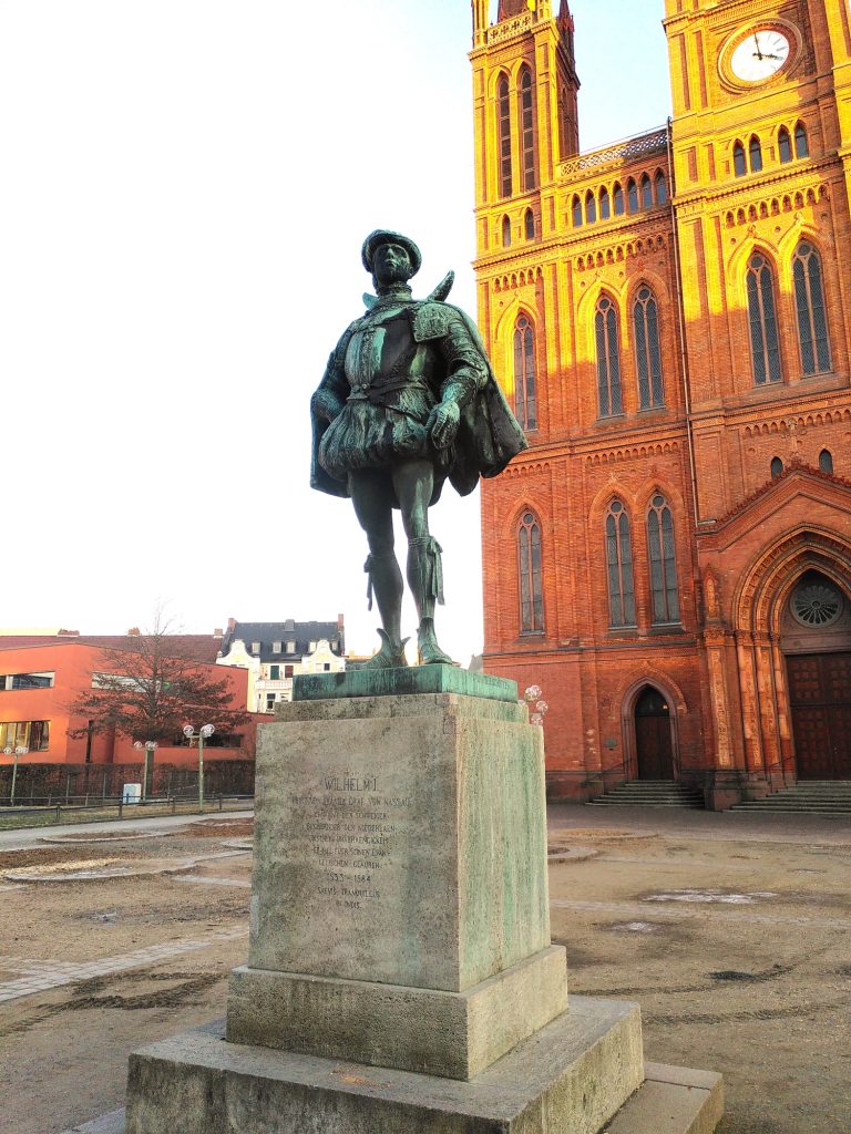 Digitale Zeitreise - Der Schweiger vor der Marktkirche Bronzestatue eines Edelmann vor einer roten Backsteinkirche.