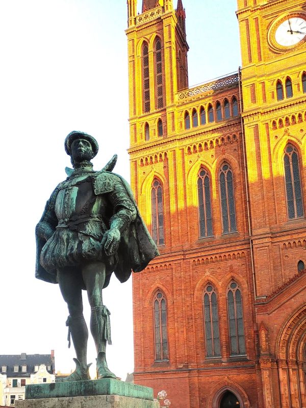 Stadtrallye Wiesbaden- Marktkirche und Wilhelm der Schweiger Bronzestatue eines Edelmanns vor einer roten Backsteinkirche mit hohen Türmen.
