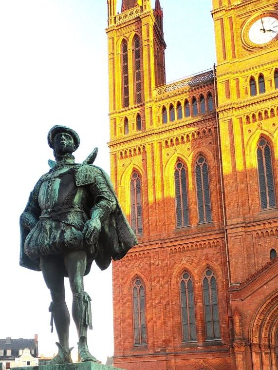 Bronzestatue eines Ritters vor einer roten Backsteinkirche mit hohen Türmen.