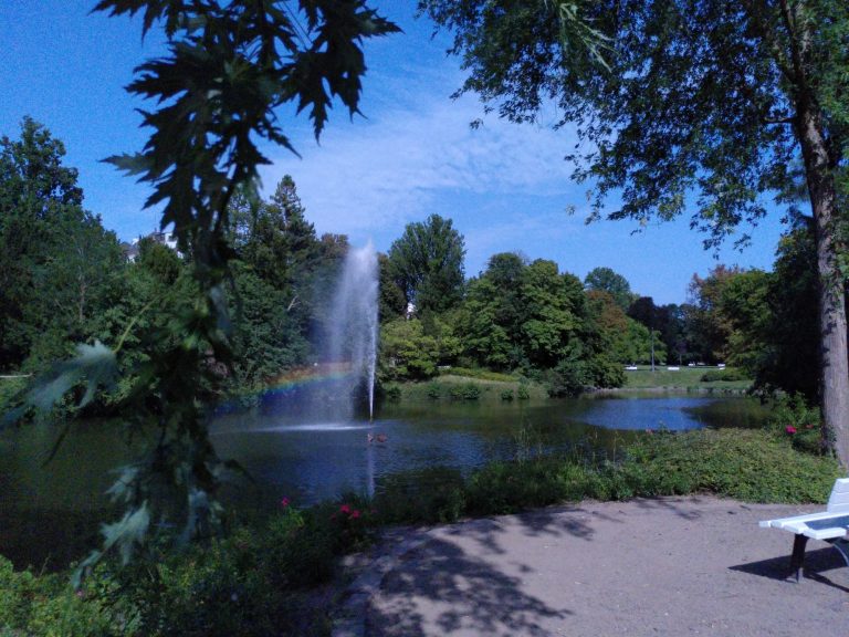 Idylle im Kurpark Ein Park mit einem Springbrunnen, einem Teich und üppigem Grün. Ein Regenbogen erscheint im Wasser.