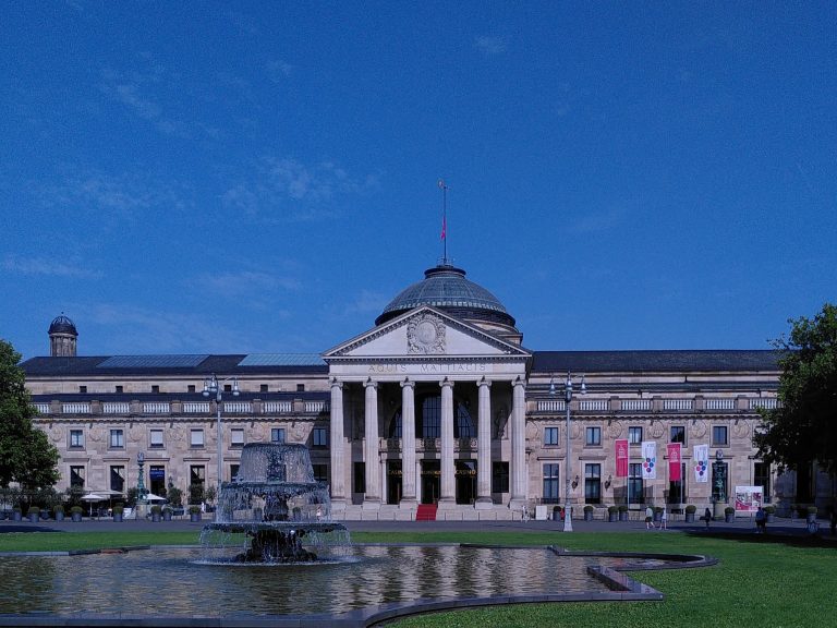 Digitale Zeitreise - Das Wiesbadener Kurhaus Historisches Gebäude mit Säulen, einem Dach und einem Brunnen im Vordergrund.