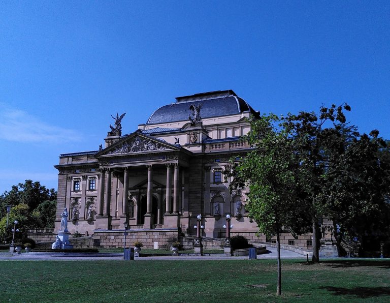 Digitale Zeitreise - Das Wiesbadener Staatstheater Historisches Gebäude mit Säulen und Skulpturen vor klarem, blauem Himmel.